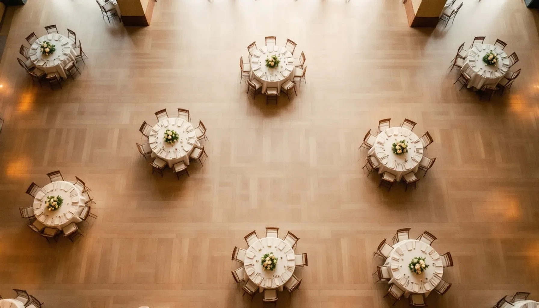 Overhead view of a wedding reception hall layout with round tables arranged on a light wooden floor, soft warm lighting
